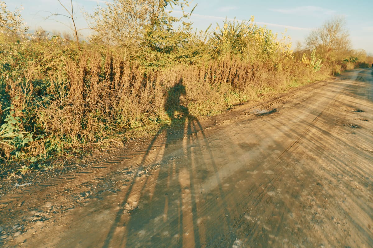 A silhouette of a cyclist riding along a countryside dirt path during sunset, creating a long shadow.